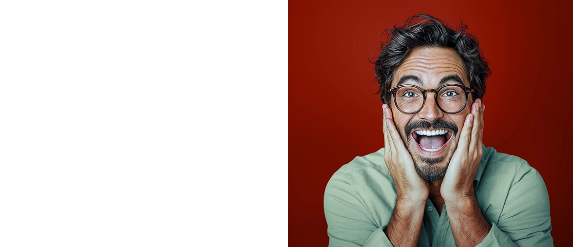 A man with black hair and dark glasses looks into the camera with his mouth open in excitement. He is wearing a light green jumper and standing in front of a dark red wall.