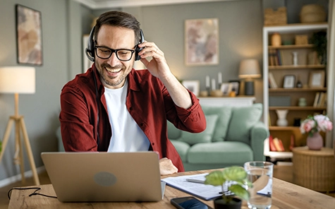 A man is working from home in his living room on his laptop and talking on the phone using a headset.