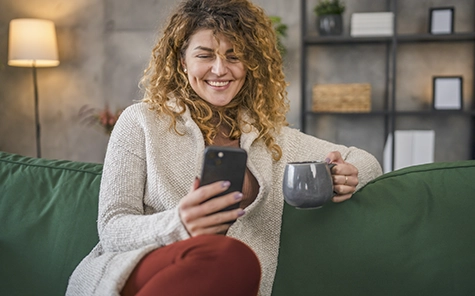 A woman sits on a cosy green sofa and uses her smartphone to control her smart home.