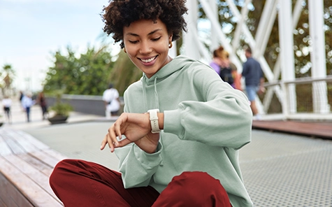 A young woman in a green jumper and red trousers sits outside on a wall and looks at her smartwatch in a good mood