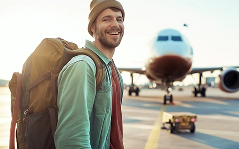 A man smiles at the camera, standing on an airfield with an aeroplane in the background. The man is wearing a knitted cap, rucksack, shirt and T-shirt.