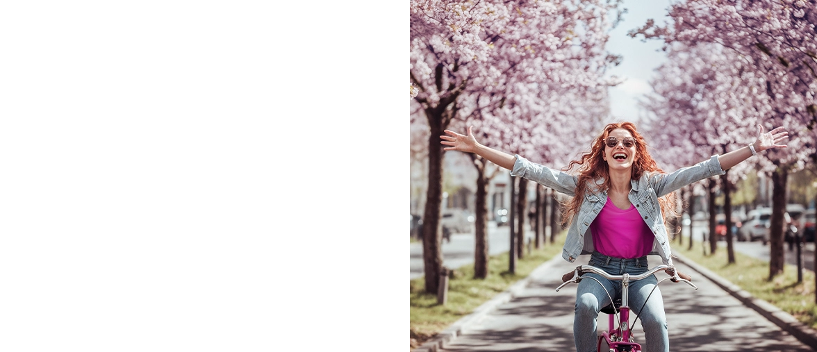 A young woman with curly red hair sits radiantly on a bicycle and cycles along an avenue adorned with cherry blossom trees with her arms outstretched.
