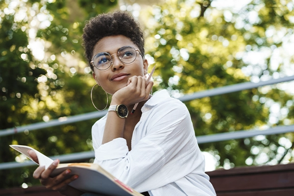 A woman in a business look sits outside on a bench under trees and gazes dreamily past the camera. She is wearing the Hama Smartwatch "7000" on her wrist