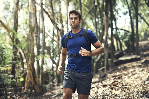 A man in a blue T-shirt and shorts jogs downhill through a forest. He is wearing the Hama Smartwatch ‘9000’ on his wrist