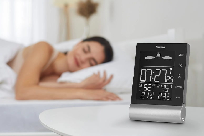 In the foreground, the Hama "Black Line" weather station stands on a bedside table; in the background, a woman lies relaxed in bed and sleeps peacefully.