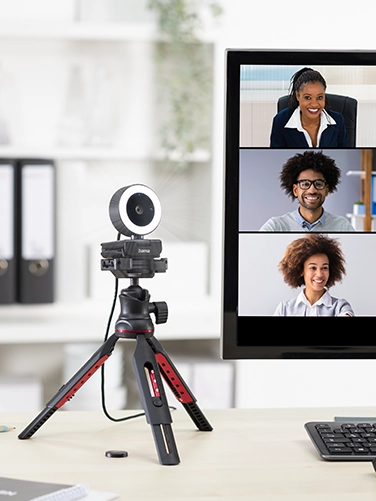 There is a Hama tripod on a desk, on which the Hama webcam ‘C-850 Pro’ with ring light is mounted. This is connected to a PC screen on which a team call with many colleagues can be seen.