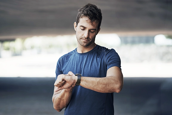 A man in a blue T-shirt stands outside under a bridge in summer weather and looks at his Hama Smartwatch ‘5000’.