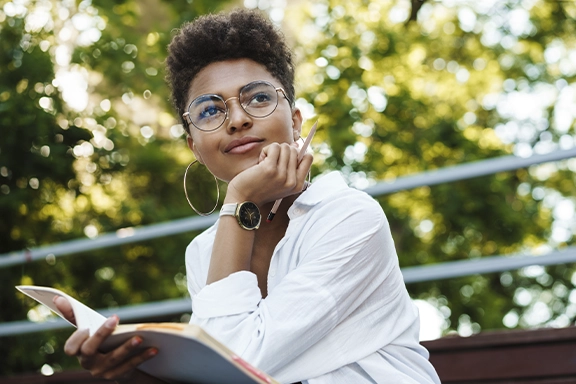 A woman in a light white blouse is sitting on a bench in the countryside. On her wrist she is wearing the Hama Smartwatch "7000" with AMOLED 1.32’ display and phone function