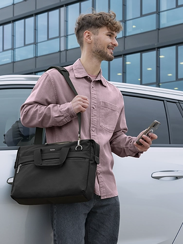 A young man leans against a silver car with the Hama ‘Ultimate’ laptop bag in black hanging over his shoulder