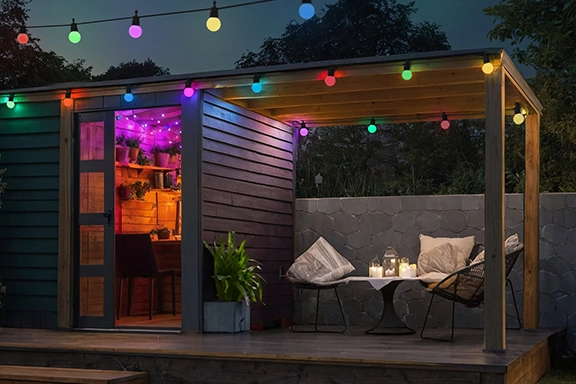 Garden scene at night in summer. In front of a cosily furnished wooden lounge, the Hama LED fairy lights bring a pleasant ambience to the garden.