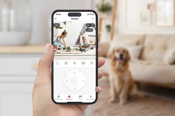 In the foreground, you can see a hand holding a smartphone with the Smart Home app open. The video transmission of the app shows two small children playing. A living room with a dog can be seen in the background of the image.