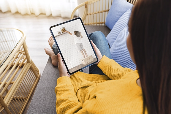 A woman in a yellow corduroy shirt looks at the screen of a tablet on which she is watching her toddler climb onto a wooden crate.