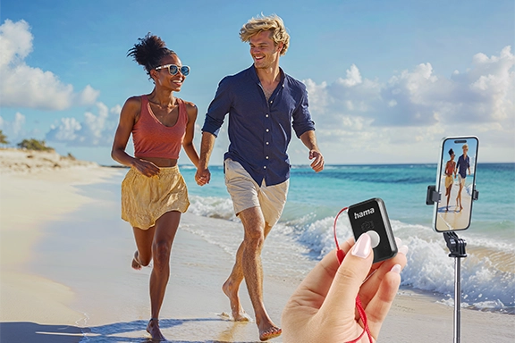 In the background, a young couple is walking along the sandy beach in a good mood and holding hands. In the foreground are cropped images of the "Fancy Stand 70 II" tripod and a hand holding the Bluetooth remote control